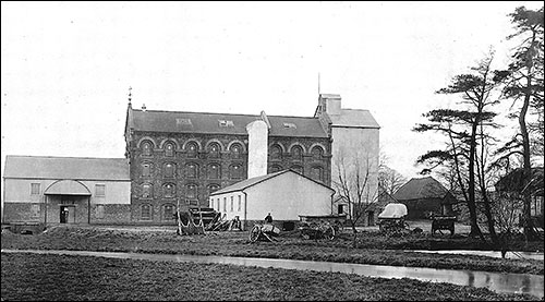 Photograph showing a selection of carts and wagons by the side of the river that passes through the mill in Station Road (early 1900s)